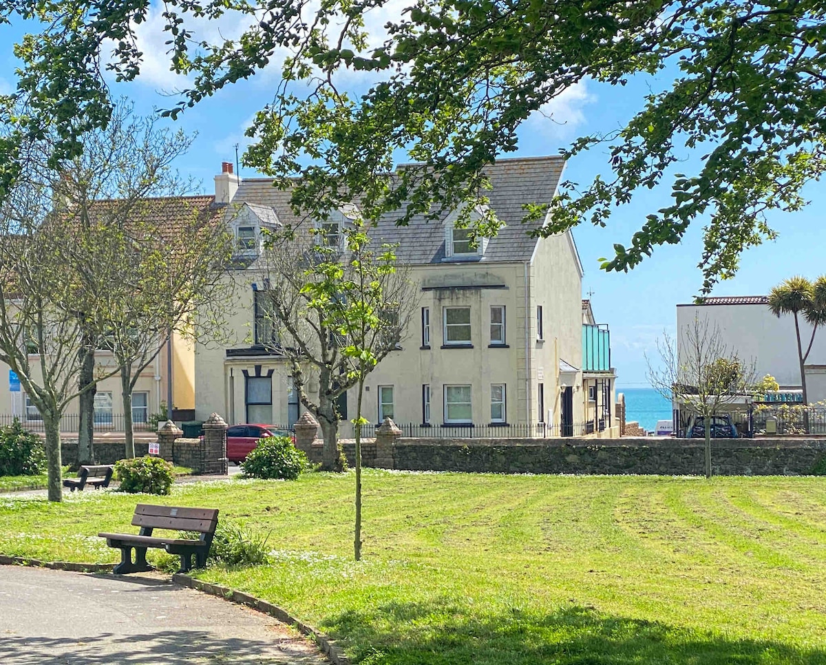 A charming view of a coastal apartment building is framed by lush greenery in a park. Soft sunlight highlights the yellow exterior of the building, while the sea glimmers in the background. A wooden bench is positioned on the manicured lawn, inviting relaxation.