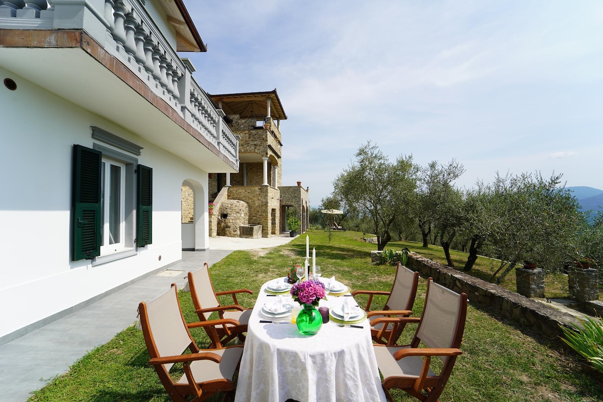 An outdoor dining area is set with a white tablecloth and elegant tableware, surrounded by six wooden chairs. The view includes expansive lawns and olive trees, with the villa's stone facade visible in the background under a clear sky.