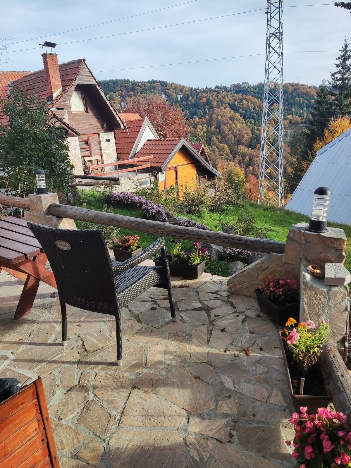 A stone patio features a wooden railing adorned with flowering pots and a comfortable chair facing a scenic view of the surrounding forested hills. Nearby, a wooden table adds to the outdoor space, enhancing the rustic atmosphere.