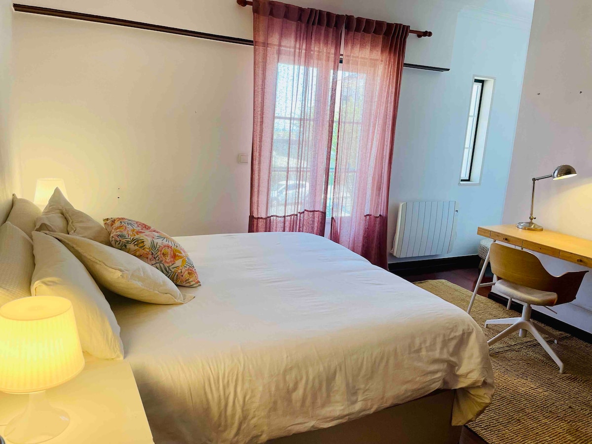 A serene bedroom is highlighted by a spacious bed adorned with decorative pillows. A warm table lamp sits on a bedside table, while a study desk with a lamp is placed near a window. Sheer curtains diffuse natural light, creating a calming atmosphere.