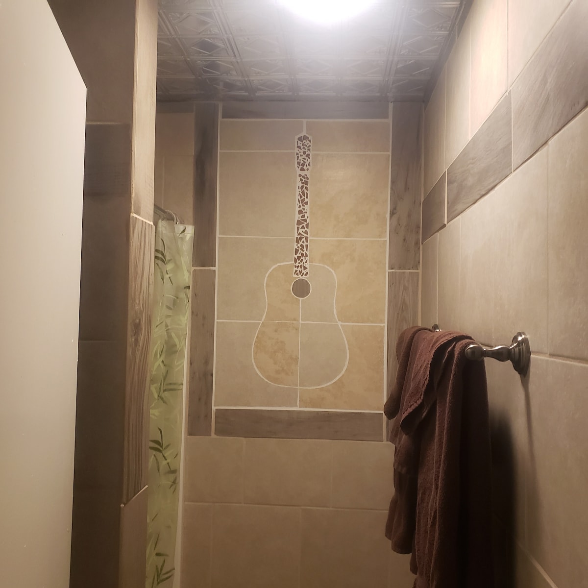 A shower area features a tiled wall adorned with a decorative guitar design, adding character to the space. A brown towel hangs on a silver towel rack, and soft lighting is provided by an overhead fixture.