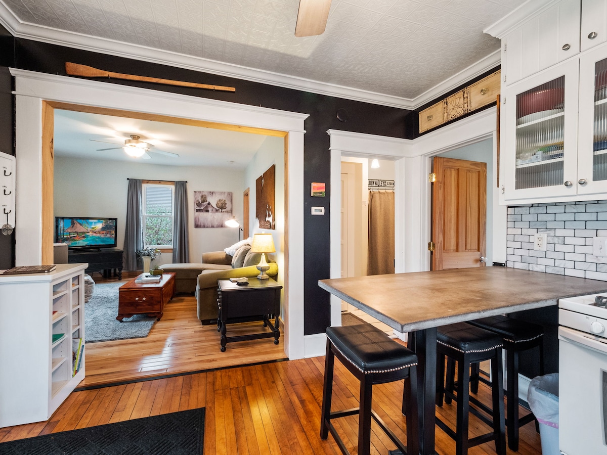 A view from the kitchen showcases a dining area with a countertop and four black chairs. The open floor plan leads into a living room with a green sofa and a large television, illuminated by natural light from a nearby window.