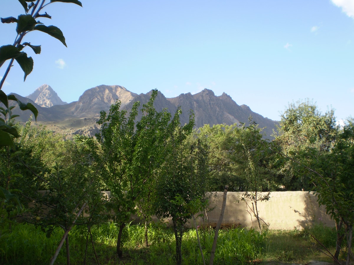 A view of surrounding hills is captured, with peaks that rise prominently against a clear blue sky. Lush greenery from fruit trees is visible in the foreground, offering a sense of tranquility and natural beauty.
