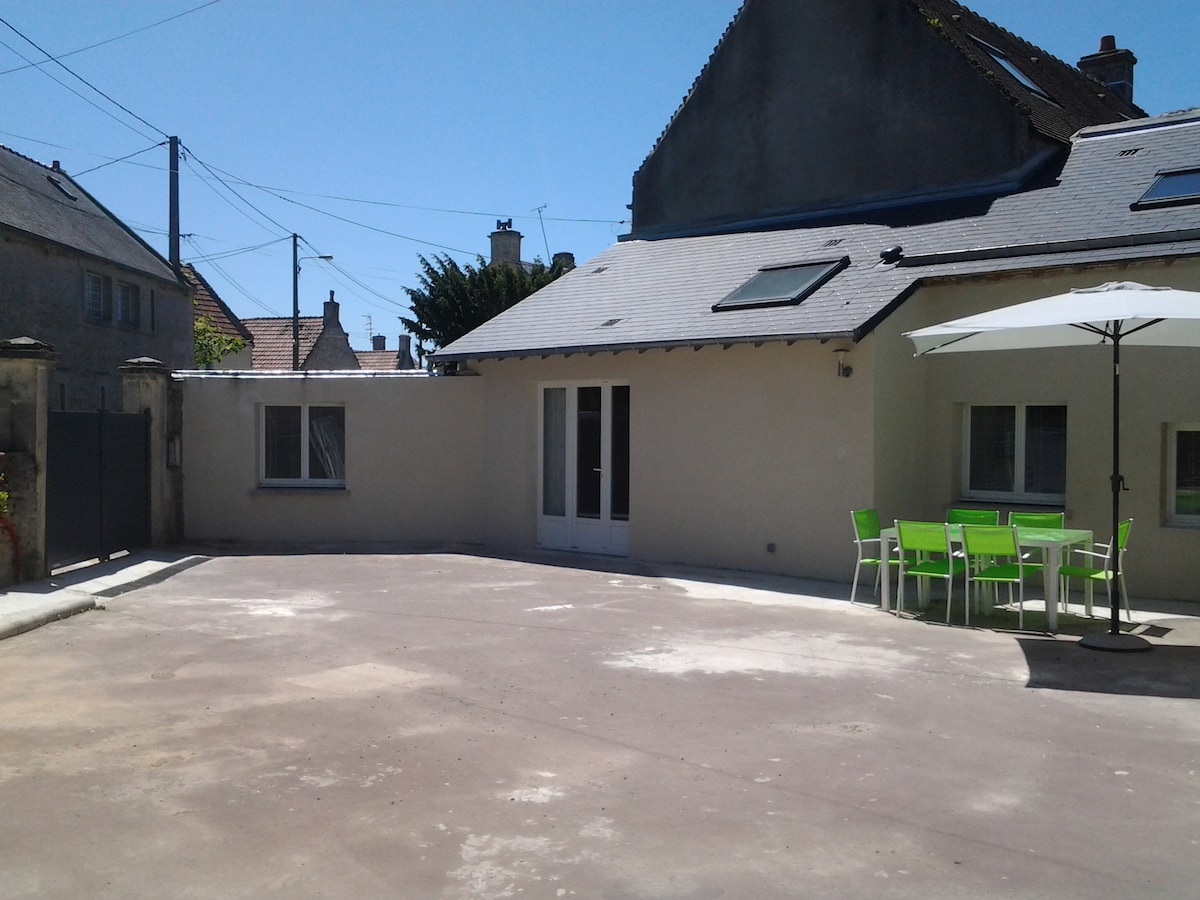 A spacious outdoor courtyard is shown, featuring a large, paved area ideal for parking. Bright green chairs surround a table under a white umbrella, providing a shaded seating option. The neighboring buildings and clear blue sky are visible in the background.
