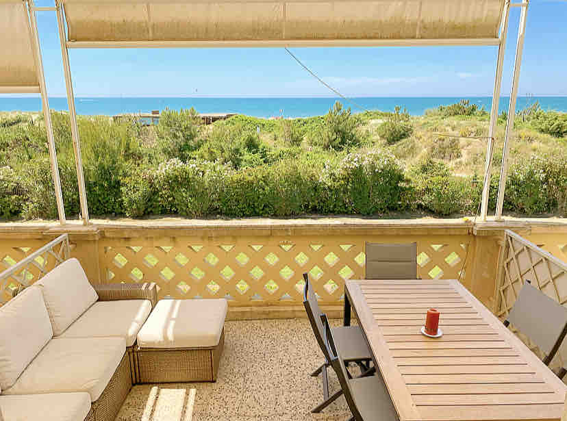 The terrace displays a comfortable seating area featuring a large sofa and plush lounge chair, surrounded by lush greenery and offering panoramic views of the sea. A dining table, set for six, includes a single decorative candle at its center, inviting relaxed outdoor meals.