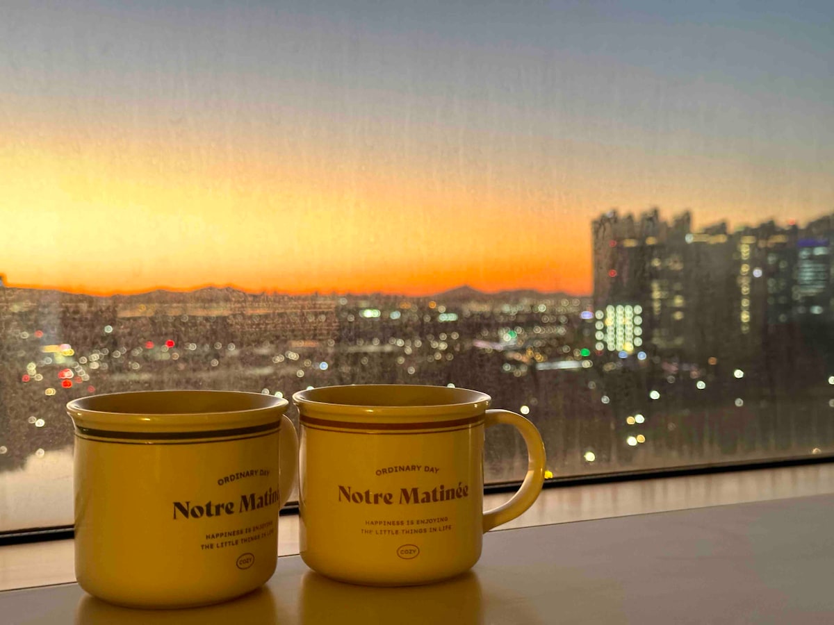 Two mugs are placed on a table, with a vibrant sunset view reflecting through the window. The city skyline is visible in the background, illuminated by the soft glow of lights as day transitions to evening.