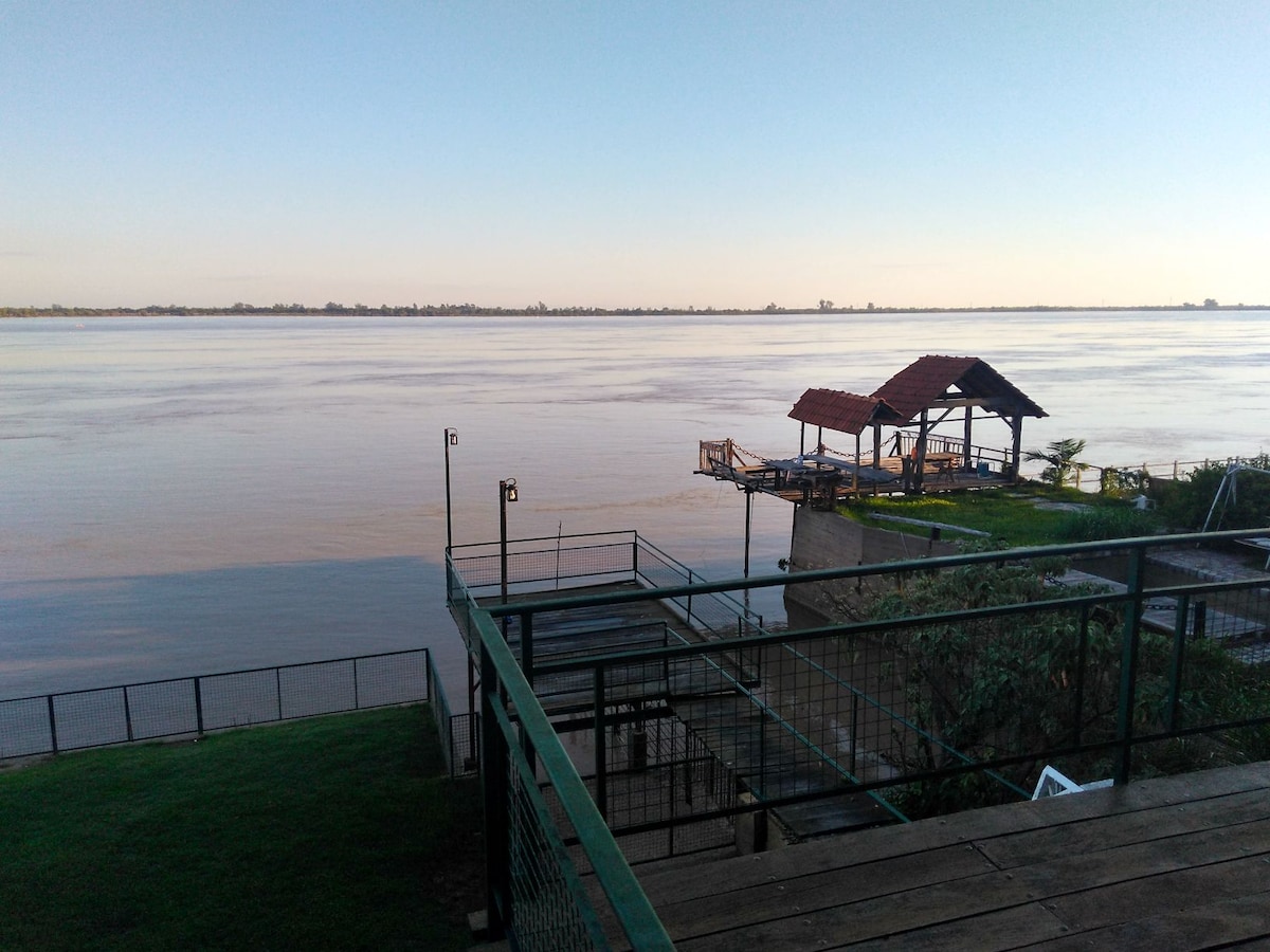 A view of the serene riverfront is shown, with calm waters extending towards the horizon. A wooden gazebo with a red roof is positioned on a dock, surrounded by green grass and a metal railing that creates a safe outdoor space.