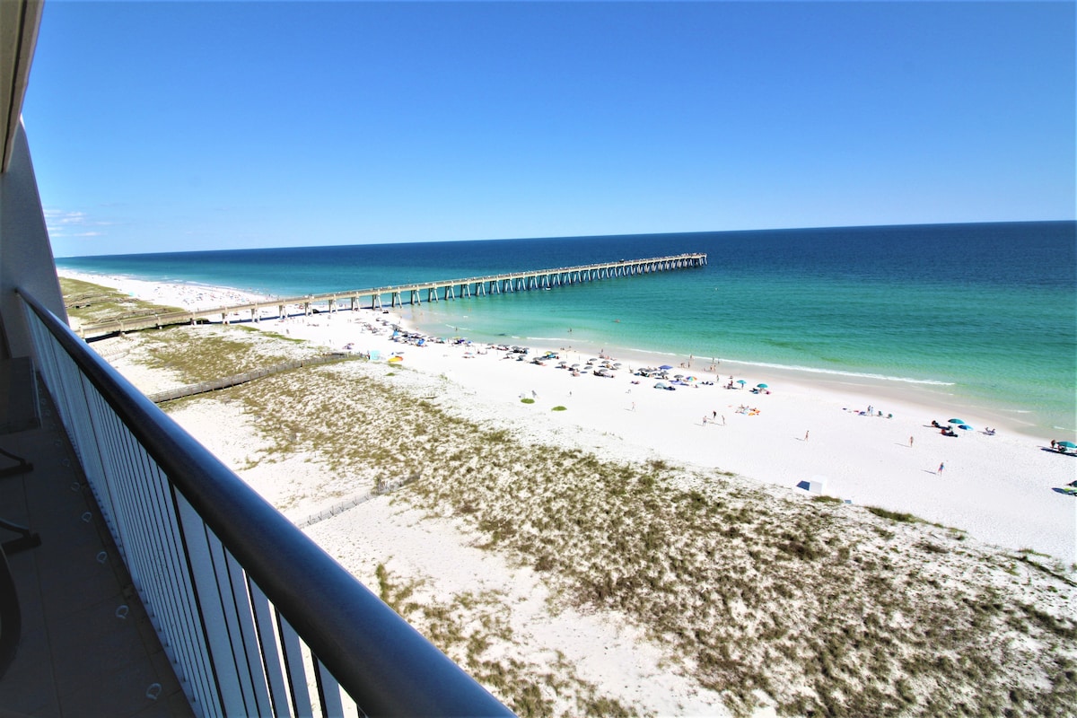 A panoramic view from the spacious balcony reveals the crystal-clear waters of the Gulf and the sandy beach below. The long fishing pier extends into the distance, complementing the serene coastal landscape bathed in natural light.
