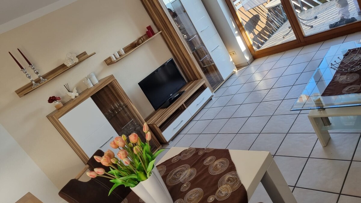 The living area features contemporary furnishings, including a low TV stand with a television and decorative wall shelves. A glass coffee table sits on the tiled floor, complemented by a dining table adorned with a vase of fresh tulips. Large windows offer natural light and views of the balcony.