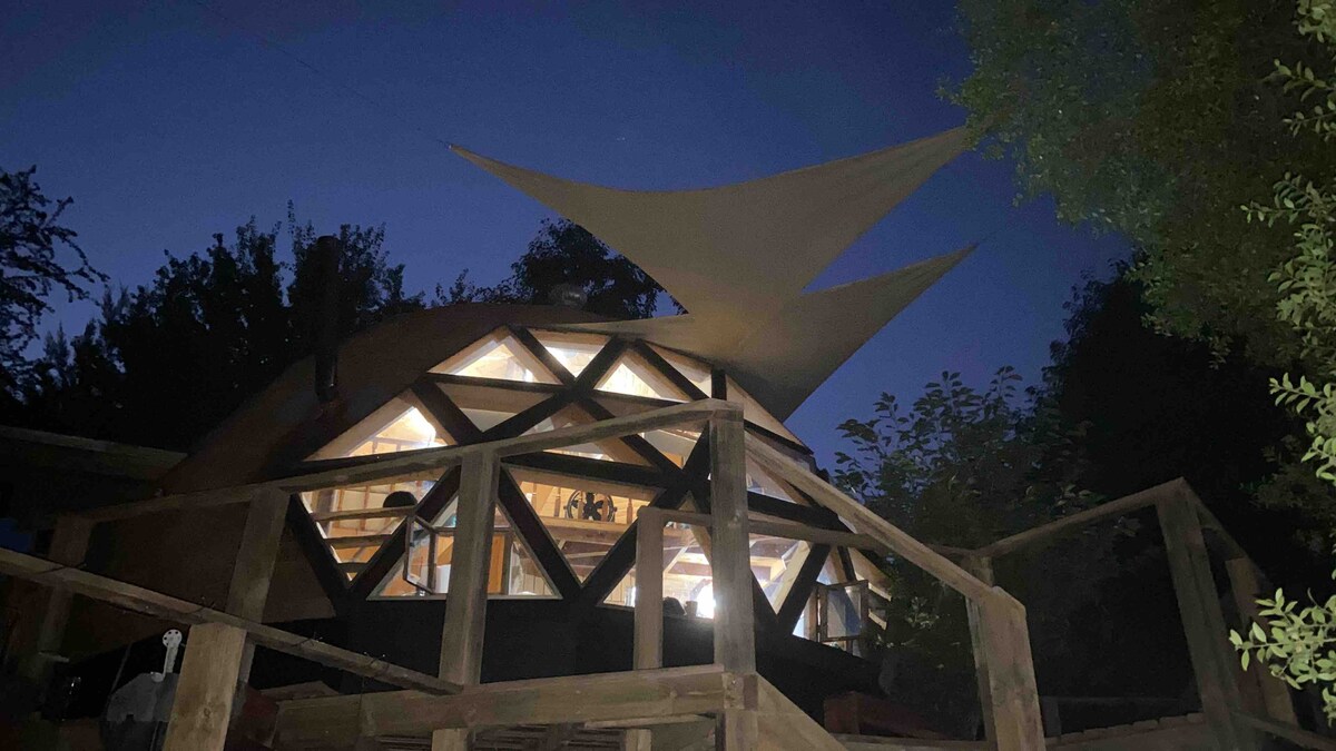 The wooden dome is illuminated at night, showcasing its unique geodesic design with triangular windows. A large fabric shade stretches above, providing additional coverage. Surrounding greenery adds to the peaceful atmosphere, while wooden decking creates an inviting entrance.