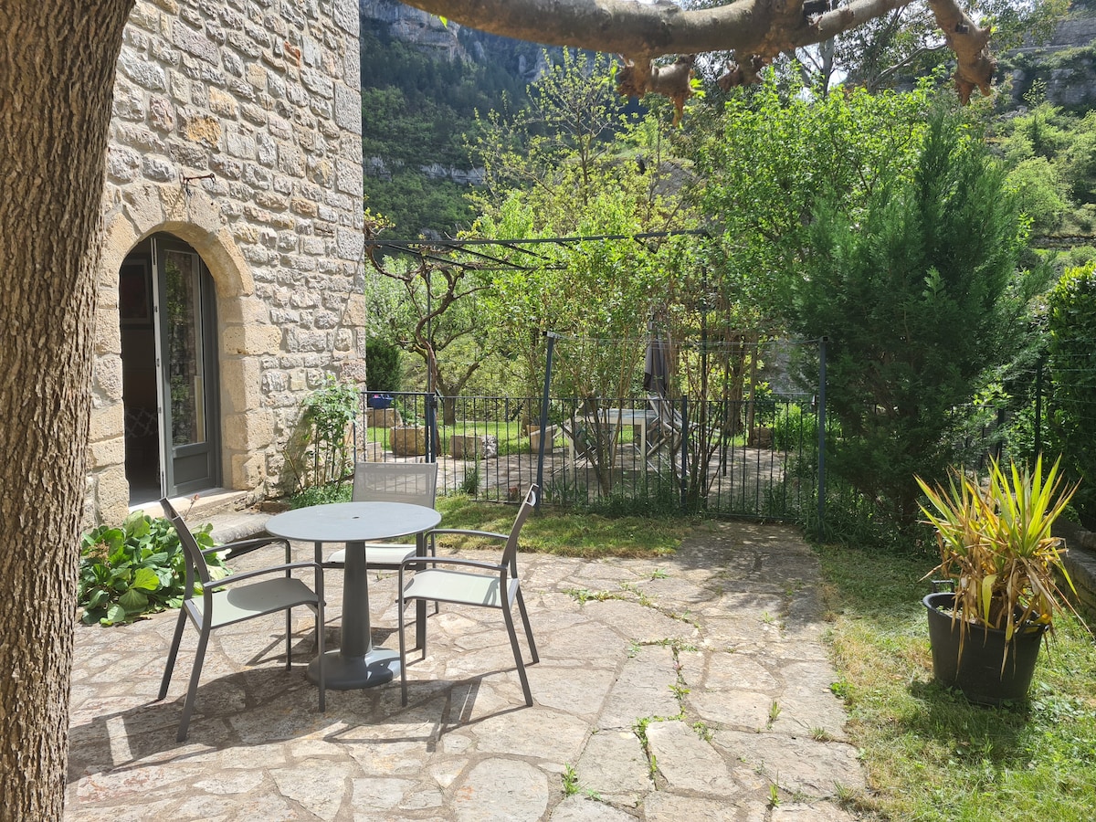 A stone patio features a round table surrounded by four chairs, set in a serene garden. Lush greenery and trees provide a tranquil backdrop, while a vine-covered arbor adds charm. The area is enclosed with a fence, ensuring privacy for guests.