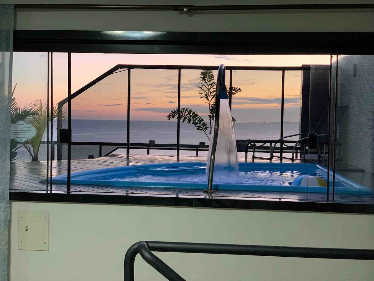 A spacious indoor area features large glass windows revealing a stunning sunset over the ocean. An inviting swimming pool can be seen in the foreground, along with outdoor seating options visible on the deck.