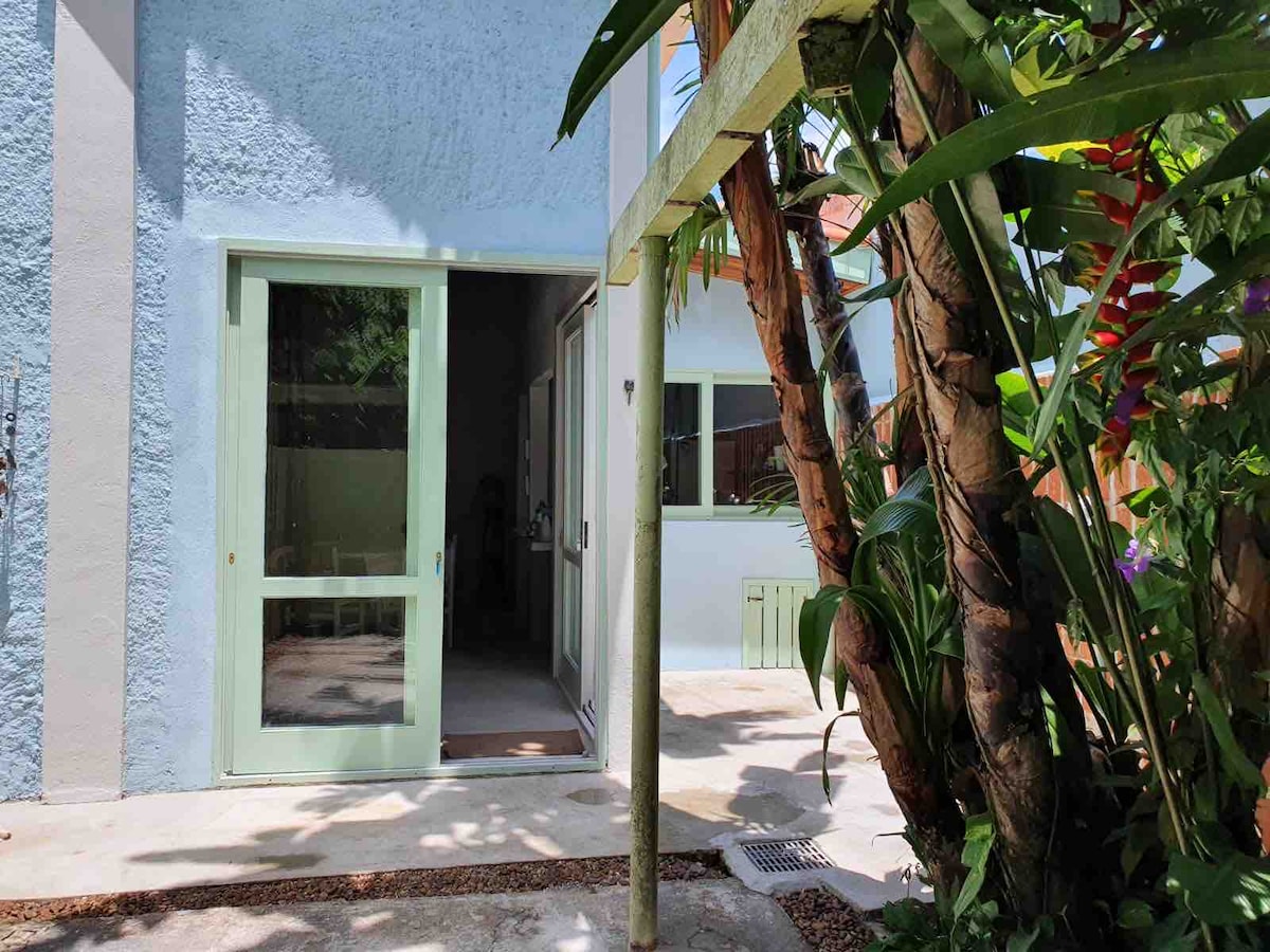An outdoor area features a light blue wall and large glass doors leading to an indoor space. Lush greenery surrounds the entrance, providing a natural ambiance. A narrow pathway with gravel is visible, enhancing the connection between the indoor and outdoor environments.