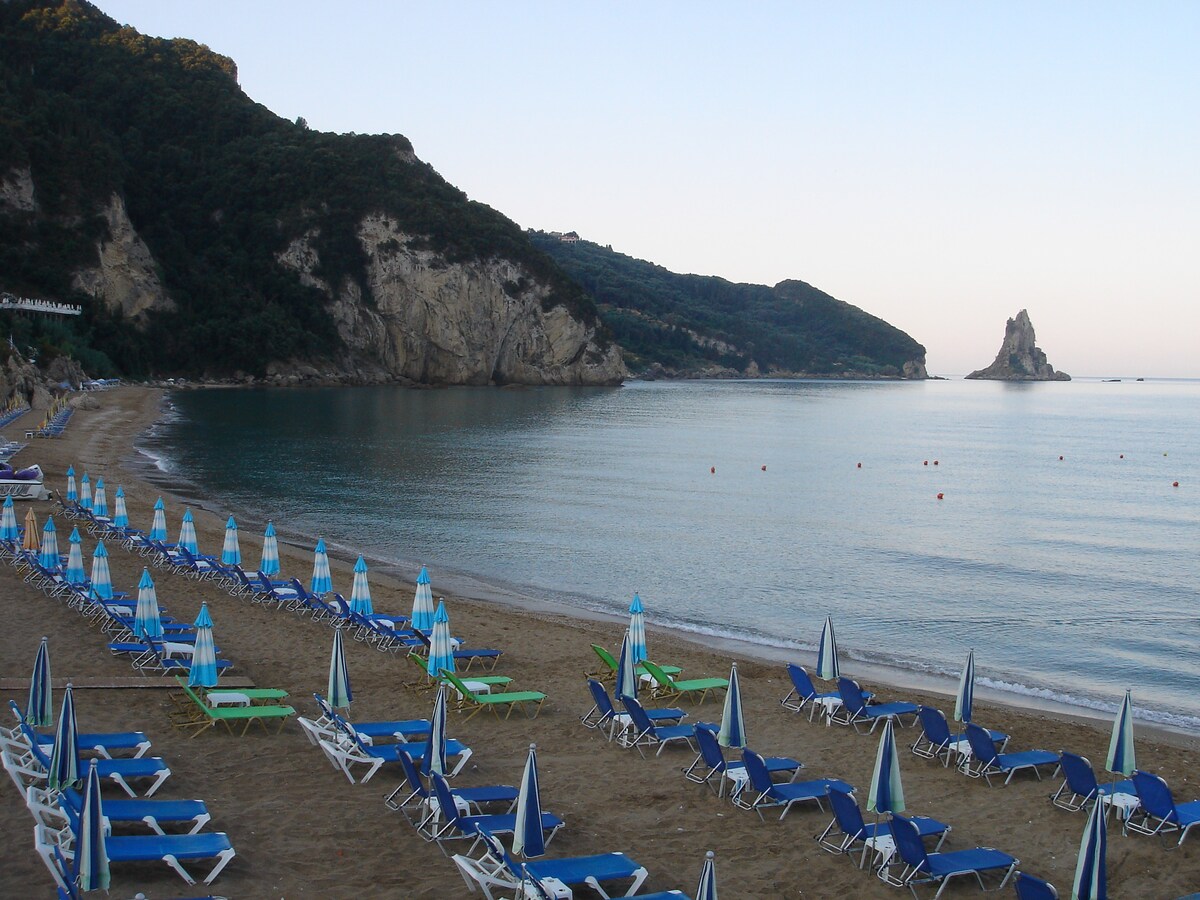 A sandy beach stretches along the shoreline, dotted with blue and white loungers under colorful striped umbrellas. The calm sea reflects the surrounding green hills and a distant rocky formation, creating a serene coastal scene. Gentle waves lap at the shore, enhancing the tranquil atmosphere.