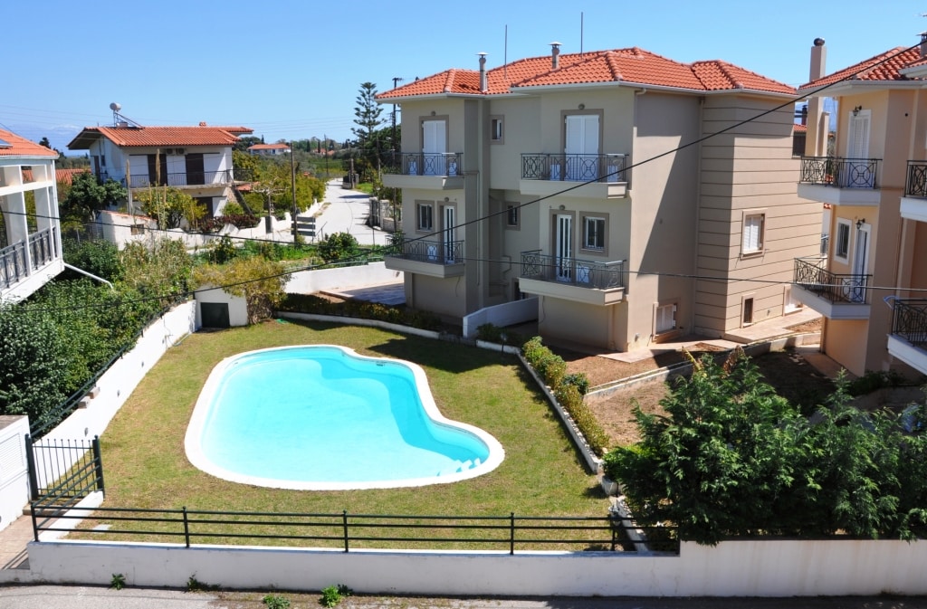 A shared swimming pool is set within a green lawn, surrounded by residential buildings. The water appears clear and inviting, while sunlit grass offers a pleasant contrast. Houses with balconies are visible in the background, contributing to a serene neighborhood atmosphere.