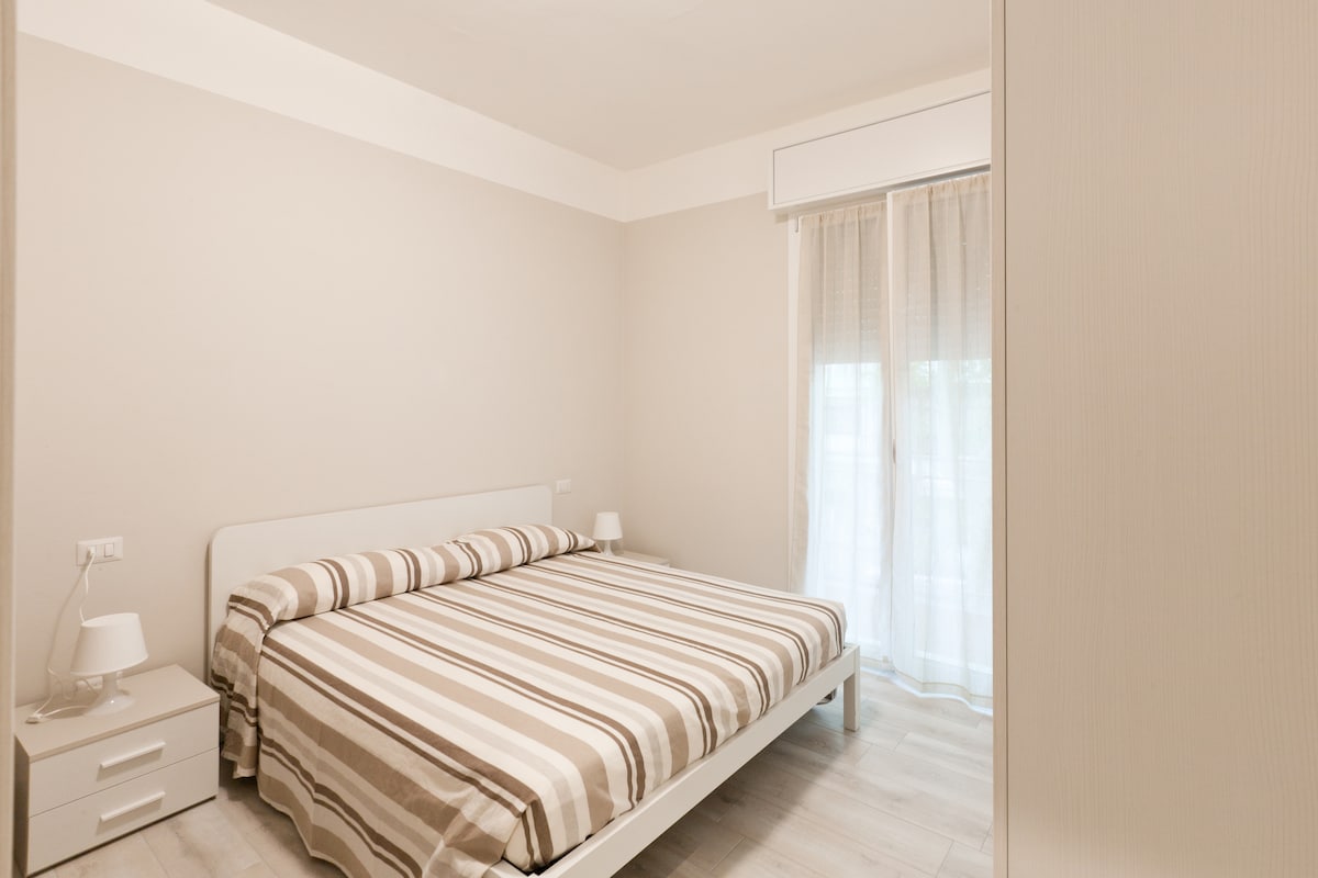 A spacious sleeping area features a striped bedspread covering a large bed, complemented by two bedside tables with lamps on either side. Natural light enters through a sheer curtain covering the window, enhancing the bright and neutral decor.