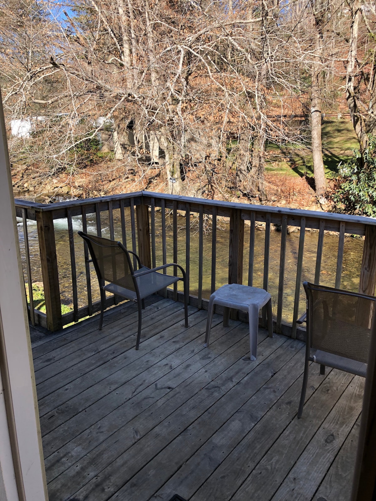 A wooden deck is featured with a railing overlooking a gently flowing river. Two metal chairs and a small table are positioned to invite outdoor relaxation. Surrounding trees are visible, showcasing natural beauty and seasonal foliage.