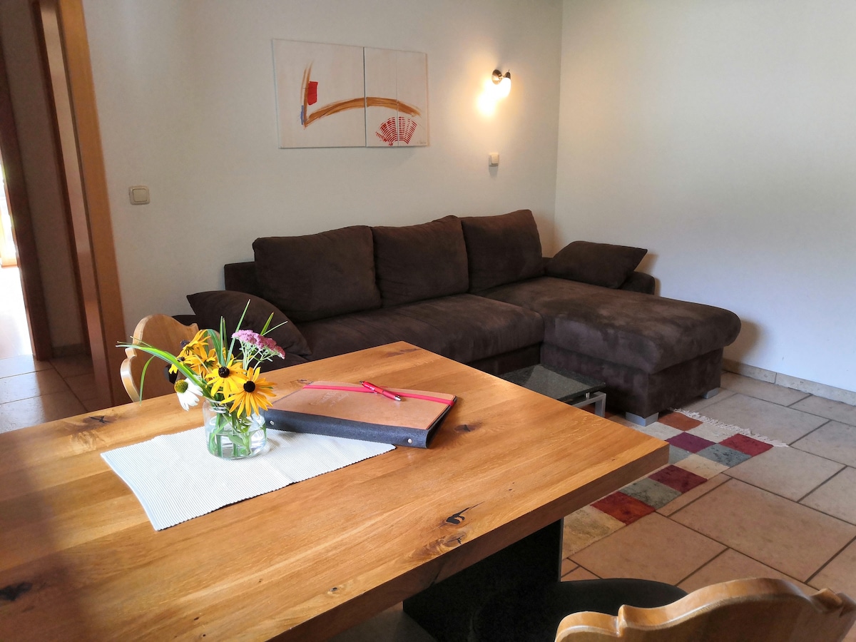 A spacious living area features a dark brown sectional sofa complemented by a wooden dining table. A small vase of flowers adds a touch of colour to the table, while light from a nearby window softly illuminates the space.