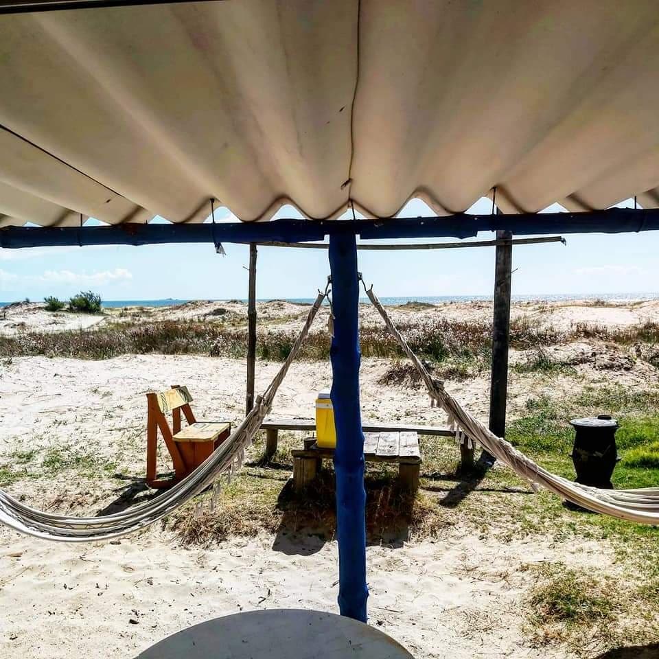 A shaded outdoor area features two hammocks hanging between sturdy posts, with a table nearby. The sandy terrain leads to a view of grassy dunes and the ocean in the distance, under a bright sky with soft clouds.