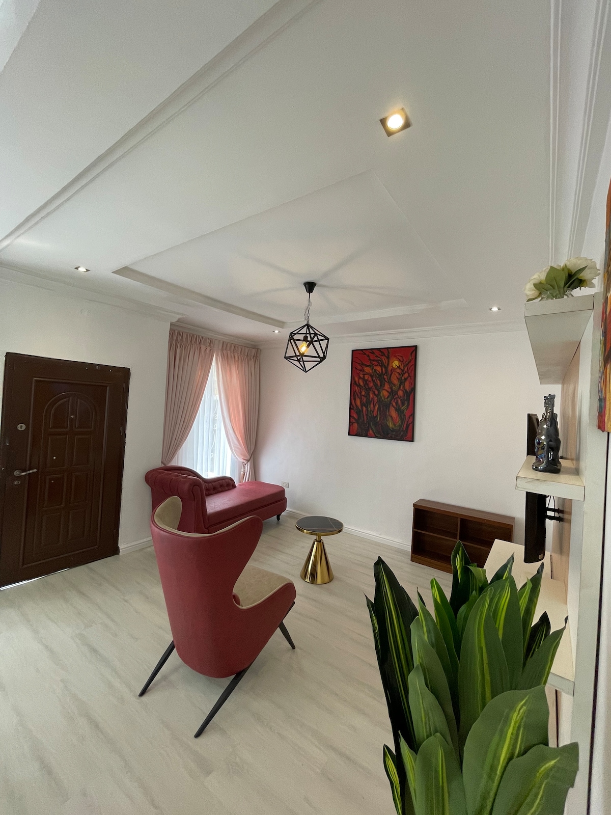 A modern sitting area features a red armchair and a small metallic side table, complemented by a stylish light fixture. A wall-mounted shelf and a framed artwork add visual interest, while lush greenery enhances the space. Natural light enters through sheer curtains.