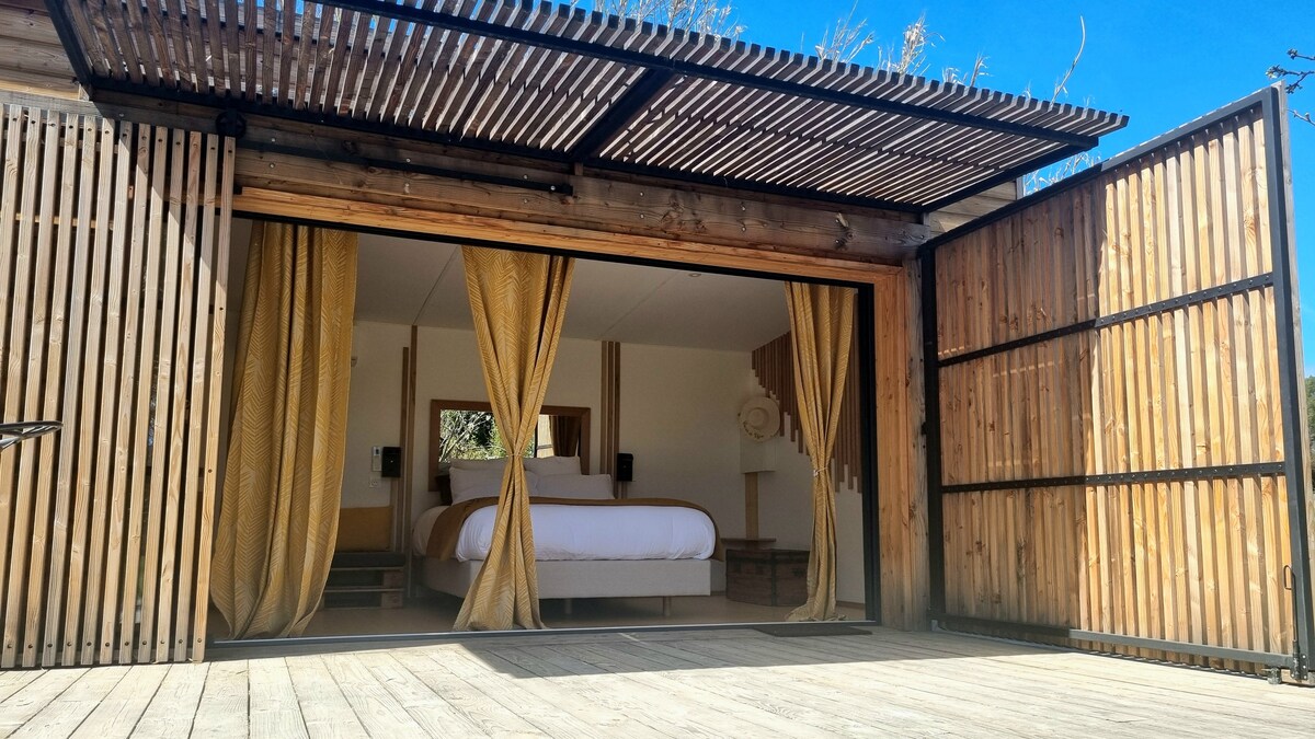 A spacious room is framed by large sliding wooden doors, which open onto a wooden deck. Soft drapes in warm hues frame the entrance, inviting natural light to illuminate the inviting sleeping area and its minimalistic furnishings.