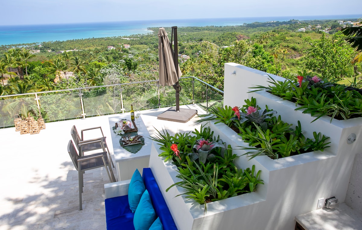 An outdoor terrace offers breathtaking views of the ocean and surrounding greenery. Potted plants and flowers are integrated with the design, while a shaded seating area features a small table and chairs. A large umbrella provides protection from the sun.