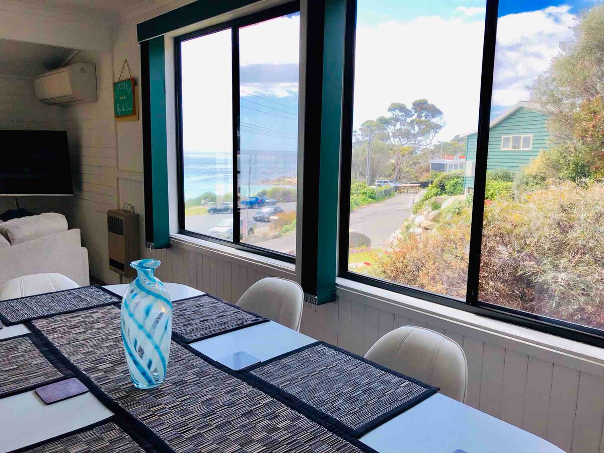 A dining table with a textured runner and decorative vase is positioned in front of large windows, providing views of the surrounding landscape and Binalong Bay. Natural light illuminates the space, highlighting the cozy interior decorated in soft, neutral tones.