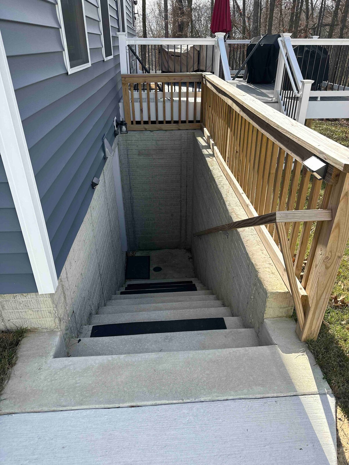 Exterior stairs lead down to the private entrance of the guest suite. The wooden railing provides safety along the steps, which are partially surrounded by a concrete wall. A grassy area is visible on one side of the staircase.