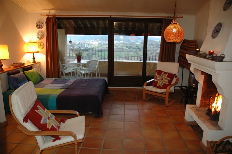 A cozy studio is featured with a spacious bed adorned in colorful linens. The area includes a small seating arrangement with two white chairs and a table, overlooking a scenic view through large glass doors. A fireplace is present, adding a touch of warmth.
