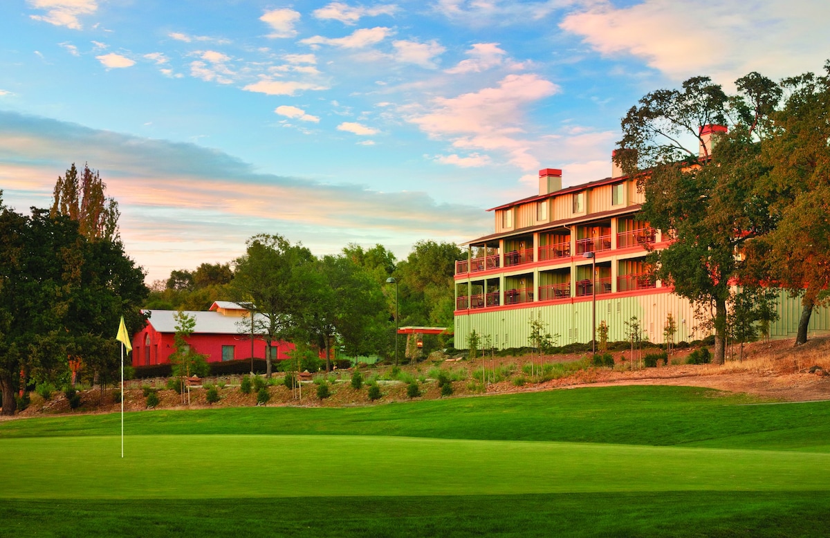 A multi-story building is nestled among green grass and trees, with a golf course flag positioned in the foreground. The building features a balcony with multiple levels, framed by vibrant landscaping and a clear sky transitioning from day to evening.