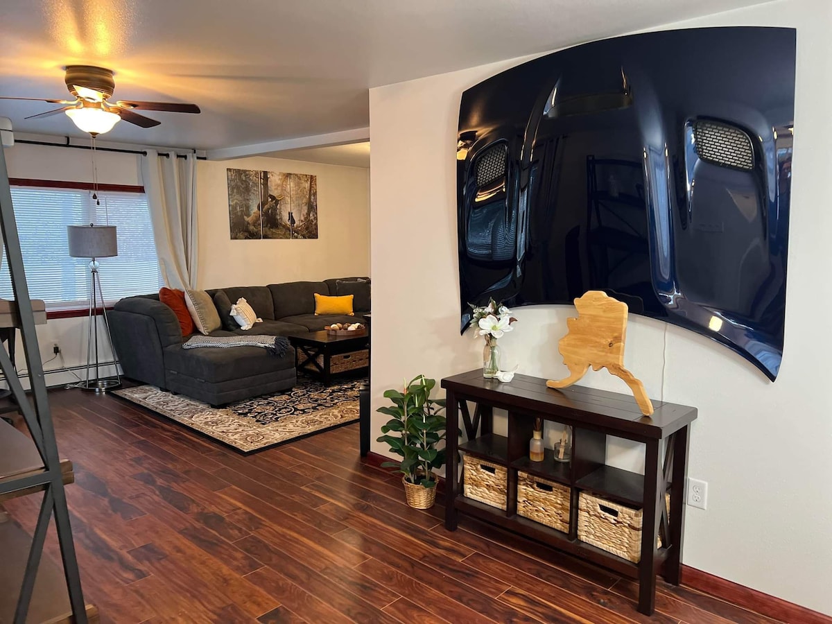 A cozy living area is presented with a large, dark sectional sofa and an area rug. An artistic piece resembling the state of Alaska is displayed on a console table, accompanied by decorative baskets and a small plant. Natural light enters through the window adorned with sheer curtains.