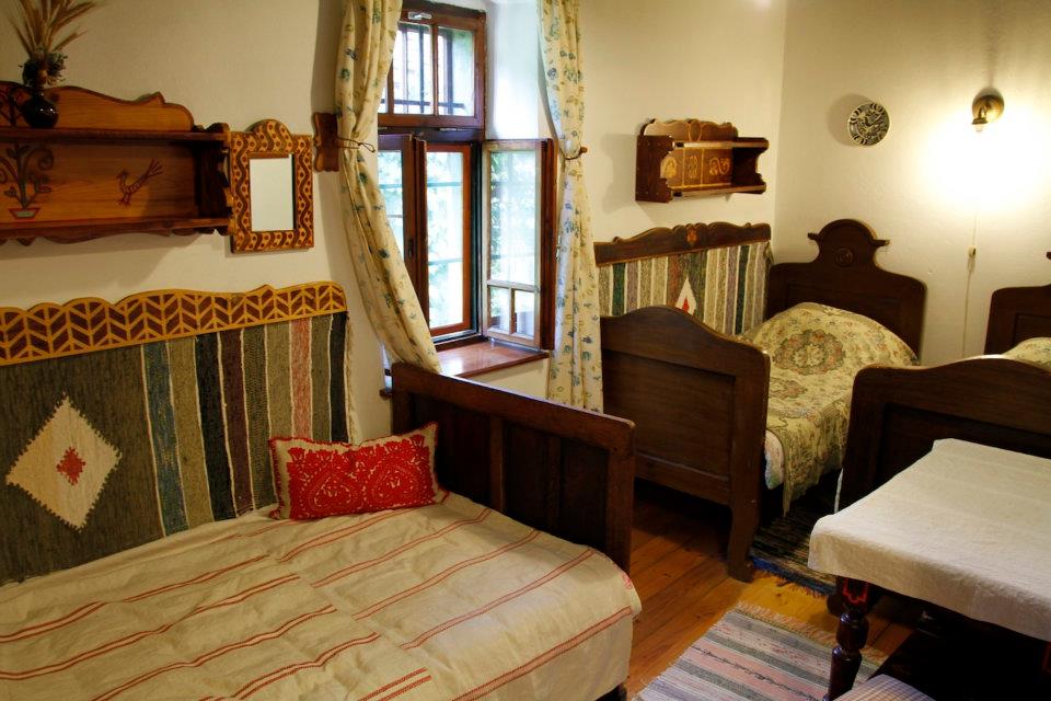 The room features three traditional wooden beds with colorful patterned headboards. A light blanket covers one of the beds. Bright windows allow natural light to fill the space, and decorative shelves hold various items, adding character to the room.