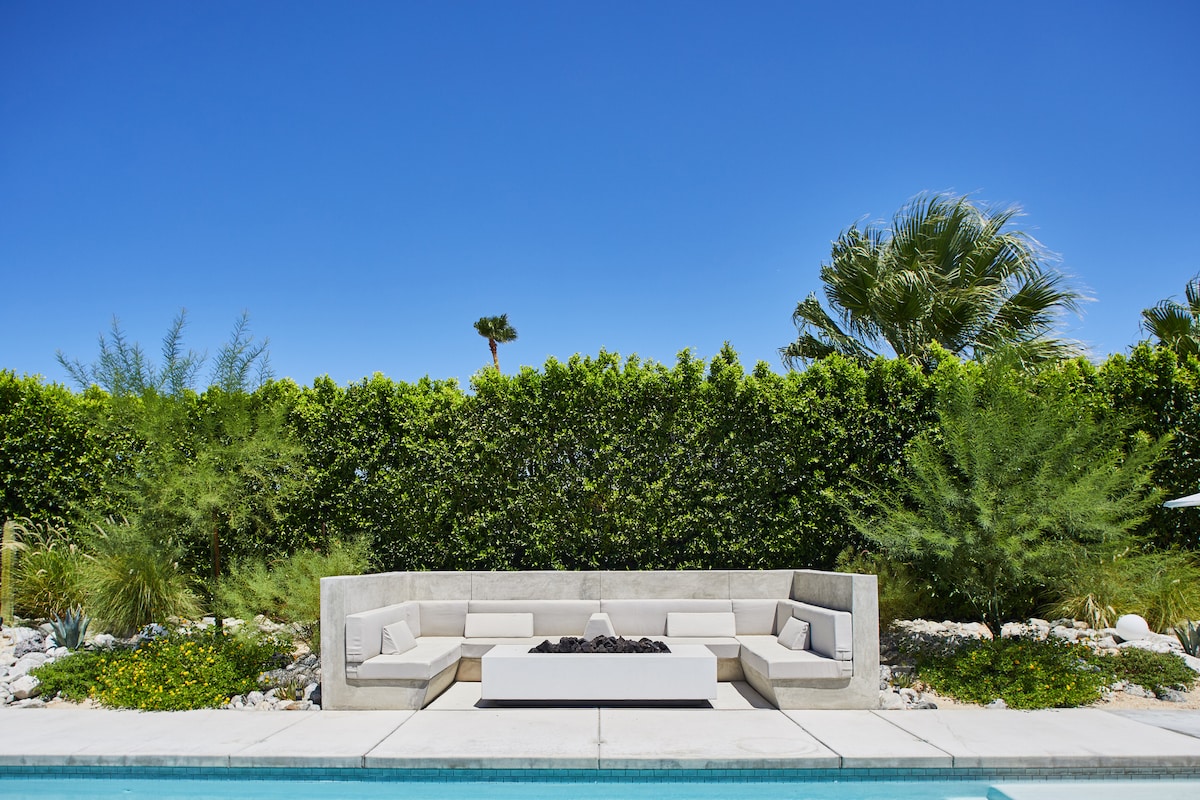 A modern fire pit area is set against a lush green hedge, featuring custom seating arranged around a rectangular fire table. The sunlit pool reflects a bright blue sky, inviting relaxation in a serene outdoor environment.