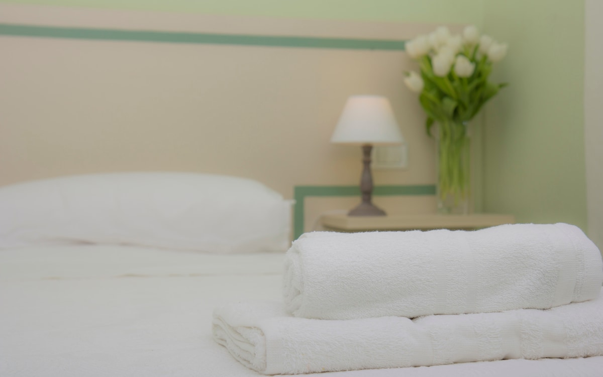 A neatly made bed is featured with fresh white linens. Two rolled towels are placed at the foot of the bed. A bedside table holds a small lamp, and a vase filled with white tulips adds a touch of color to the serene setting.