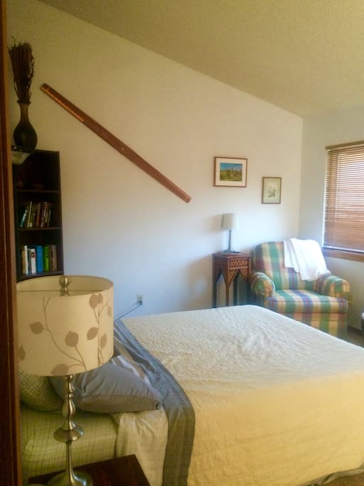 The bedroom is light and airy, with a Queen size bed, reading chair, and desk
