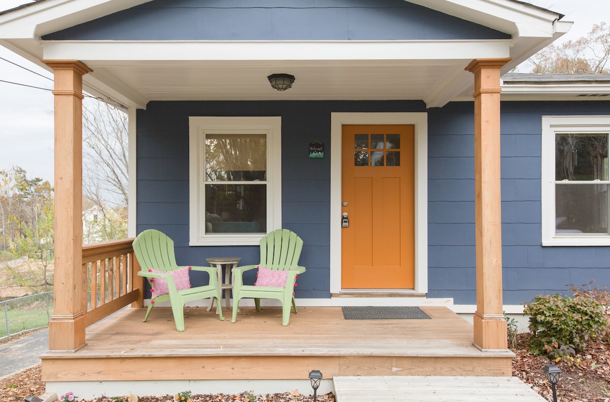 A welcoming front porch features two green chairs flanking a small table, adding a relaxed seating area. The bright orange front door creates an inviting entrance. Natural light filters through the windows, and wooden posts frame the porch, enhancing the cozy outdoor space.