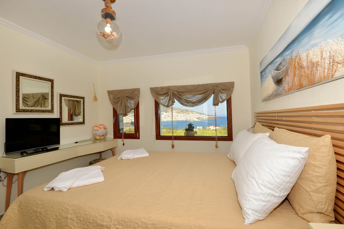 A comfortable sleeping area features a spacious bed with neutral linens and two white towels neatly placed. Large windows provide a view of the sea and surrounding landscape, while a television and a desk contribute to the room's functionality.