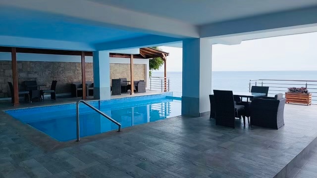 An infinity pool is viewed against a backdrop of the ocean, with seating arranged nearby. Covered areas feature tables and chairs, providing options for relaxation while enjoying the scenic waterfront. Large openings allow for natural light and breeze to flow through the space.
