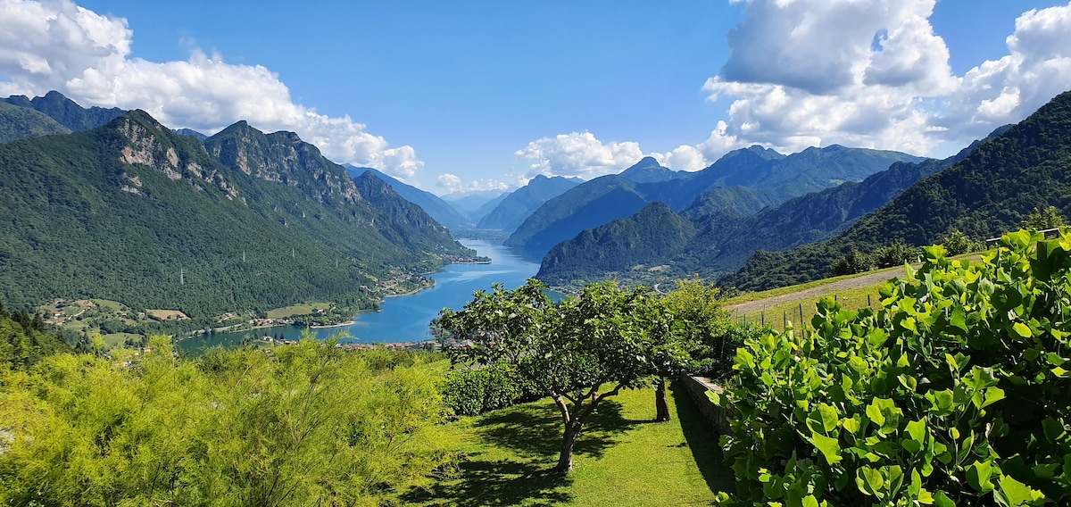 A scenic view of a lake is framed by mountainous terrain. Lush greenery fills the foreground, while the calm water reflects the surrounding peaks and sky. The landscape is open and inviting, showcasing the natural beauty of the area.