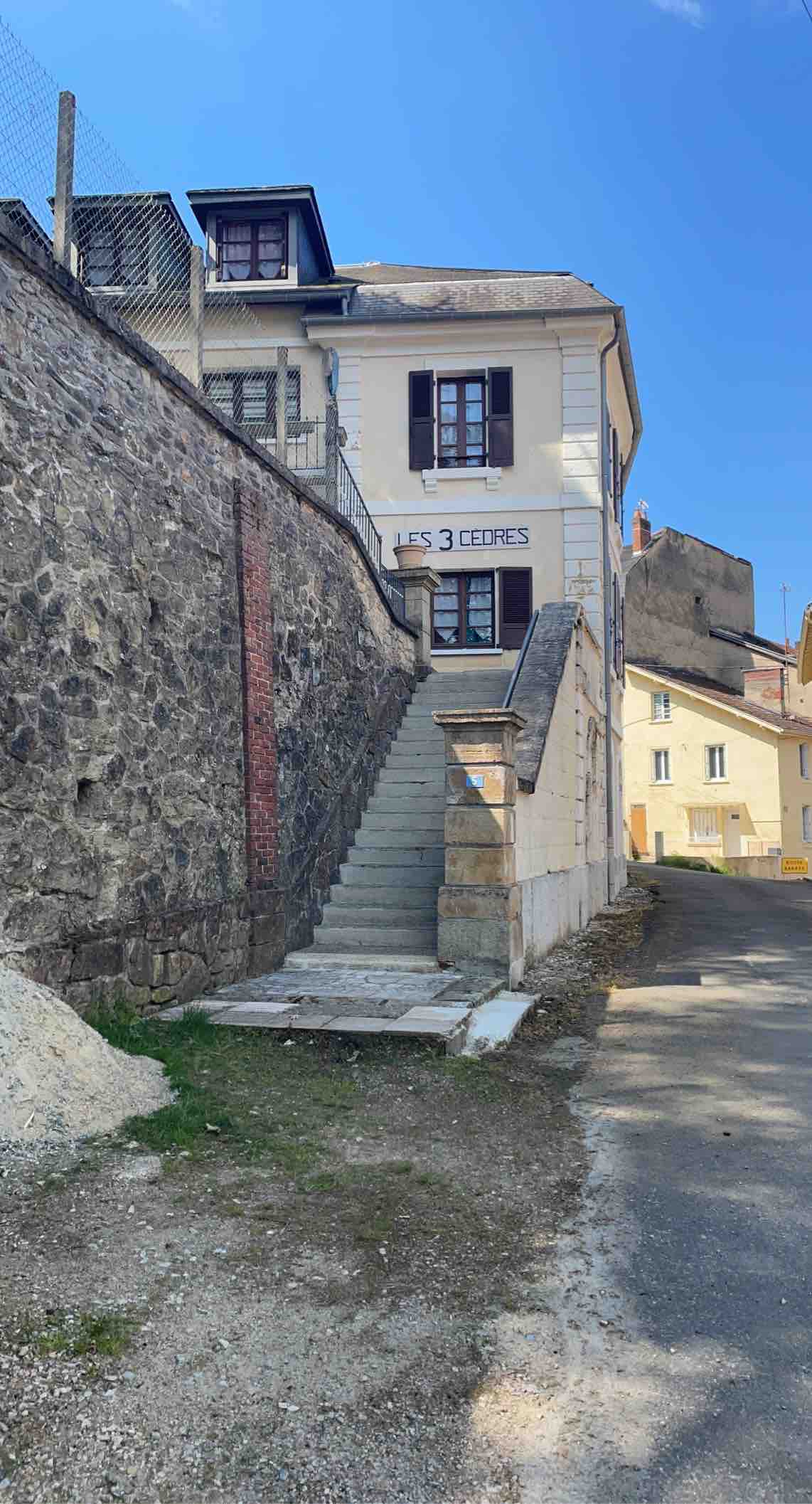 An exterior view of a building is shown, featuring a set of stone steps leading up to a second-floor entrance. The building's facade includes decorative shutters and a sign that reads 'LES 3 CEDRES.' Surrounding structures and a gravel path are visible in the background.