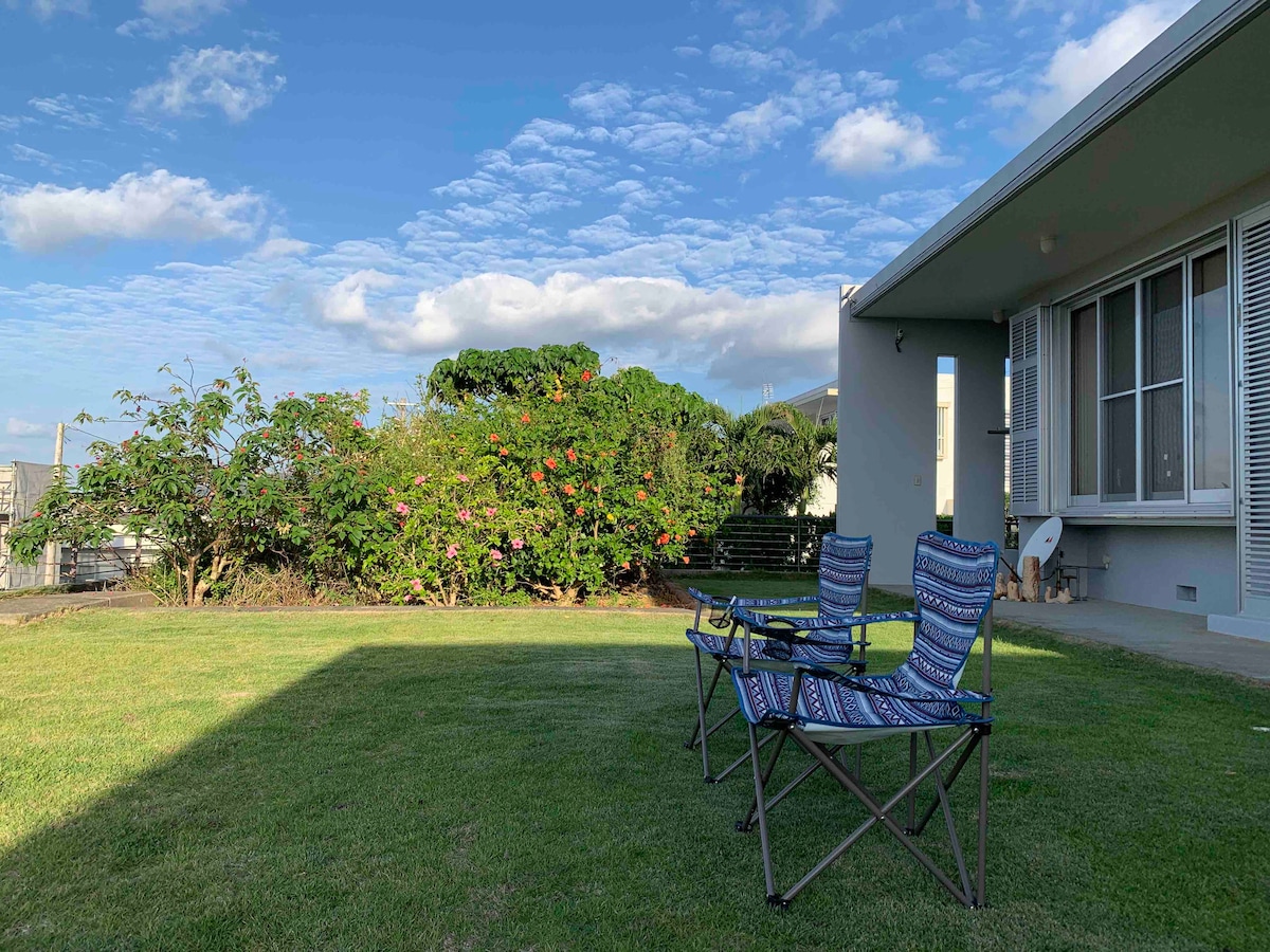 Two folding chairs are positioned on a grassy area, overlooking a well-maintained garden adorned with shrubs and flowering plants. The sky is clear with scattered clouds, creating a bright and open atmosphere in the outdoor space.