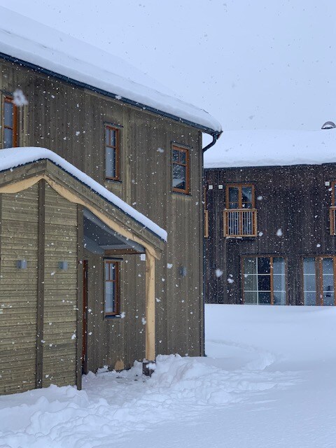 The exterior of the cabin is showcased amid falling snow, with a combination of wooden textures and muted colors. Snow blankets the ground and roof, while the nearby structure features a balcony and large windows, providing a glimpse of the snowy surroundings.