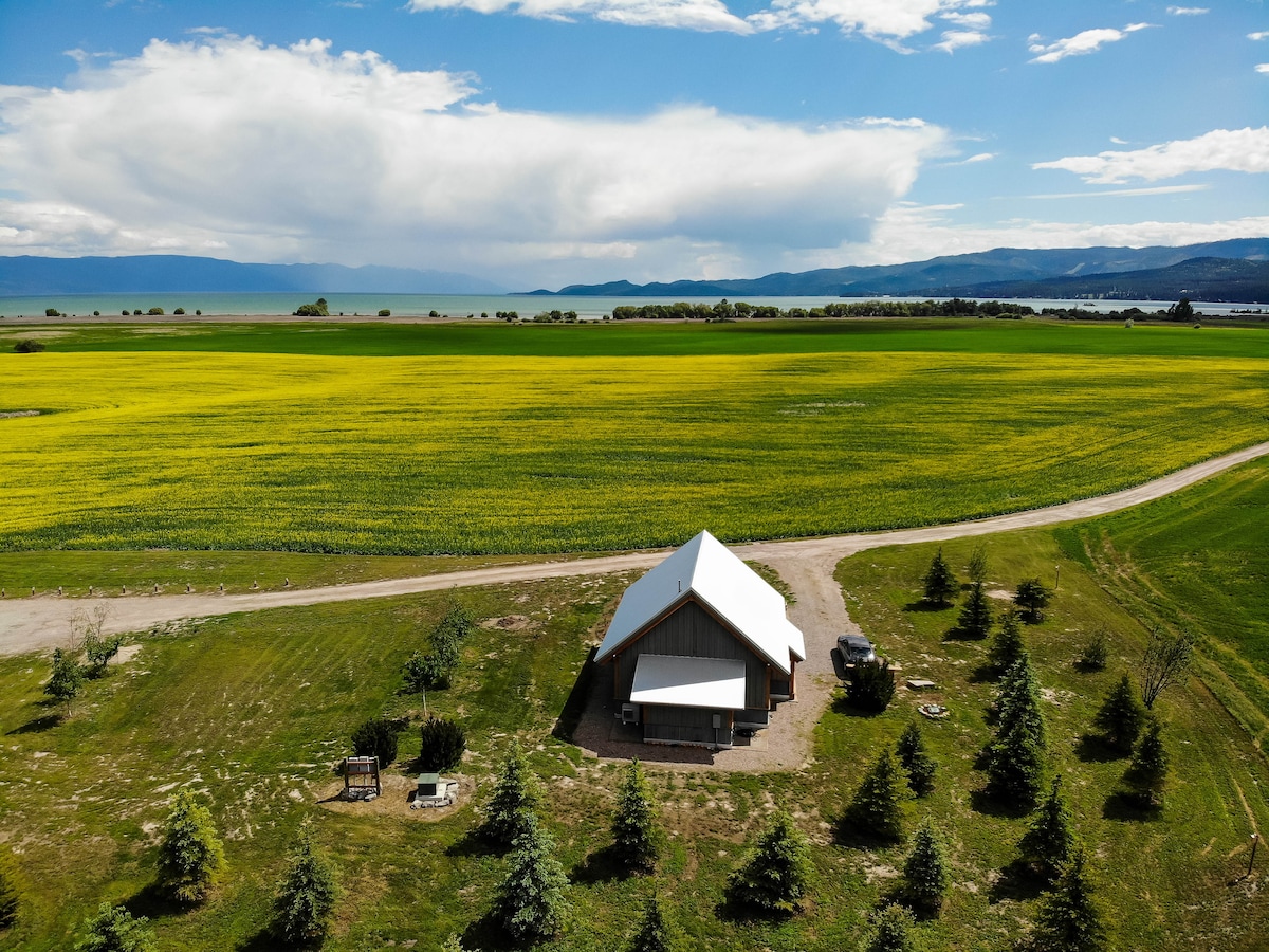 A renovated barn is situated on a vibrant green field, surrounded by an expansive landscape. The view captures the countryside leading to the distant Flathead Lake, with rolling hills and mountains visible under a wide blue sky adorned with light clouds.