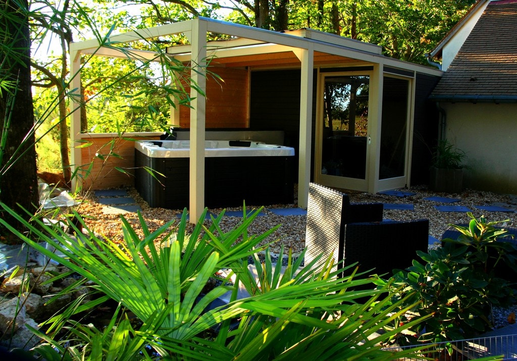 A serene outdoor area features a sheltered relaxation space with a hot tub surrounded by greenery. Comfortable seating arrangements are provided, along with pebbled pathways and surrounding plants that enhance the tranquil ambiance.