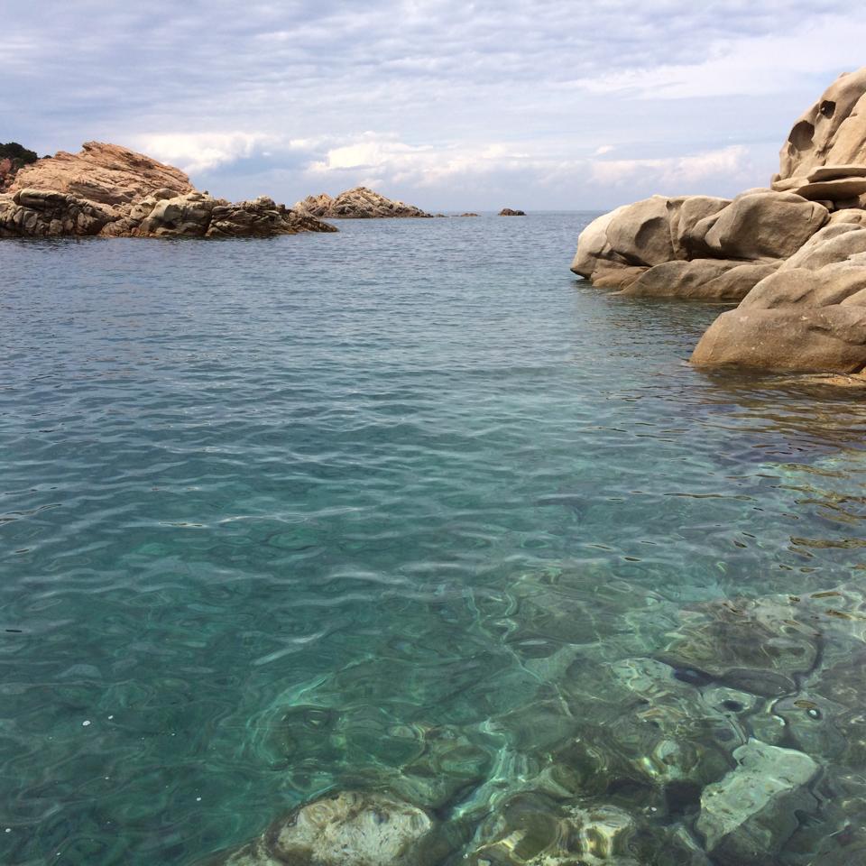Clear turquoise waters reveal a rocky shoreline, with natural formations jutting above the surface. The calm sea reflects a soft sky, showcasing a serene coastal environment. The visibility of submerged rocks emphasizes the marine landscape's beauty.