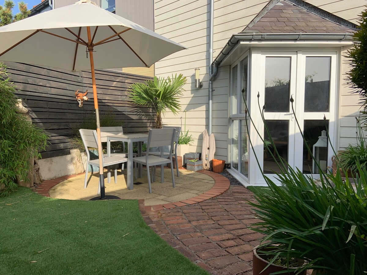 A private garden area features a round seating arrangement with a table and four chairs, shaded by a large umbrella. Decorative plants add greenery, while a brick path leads to a corner with a glass door, providing access to the cottage.