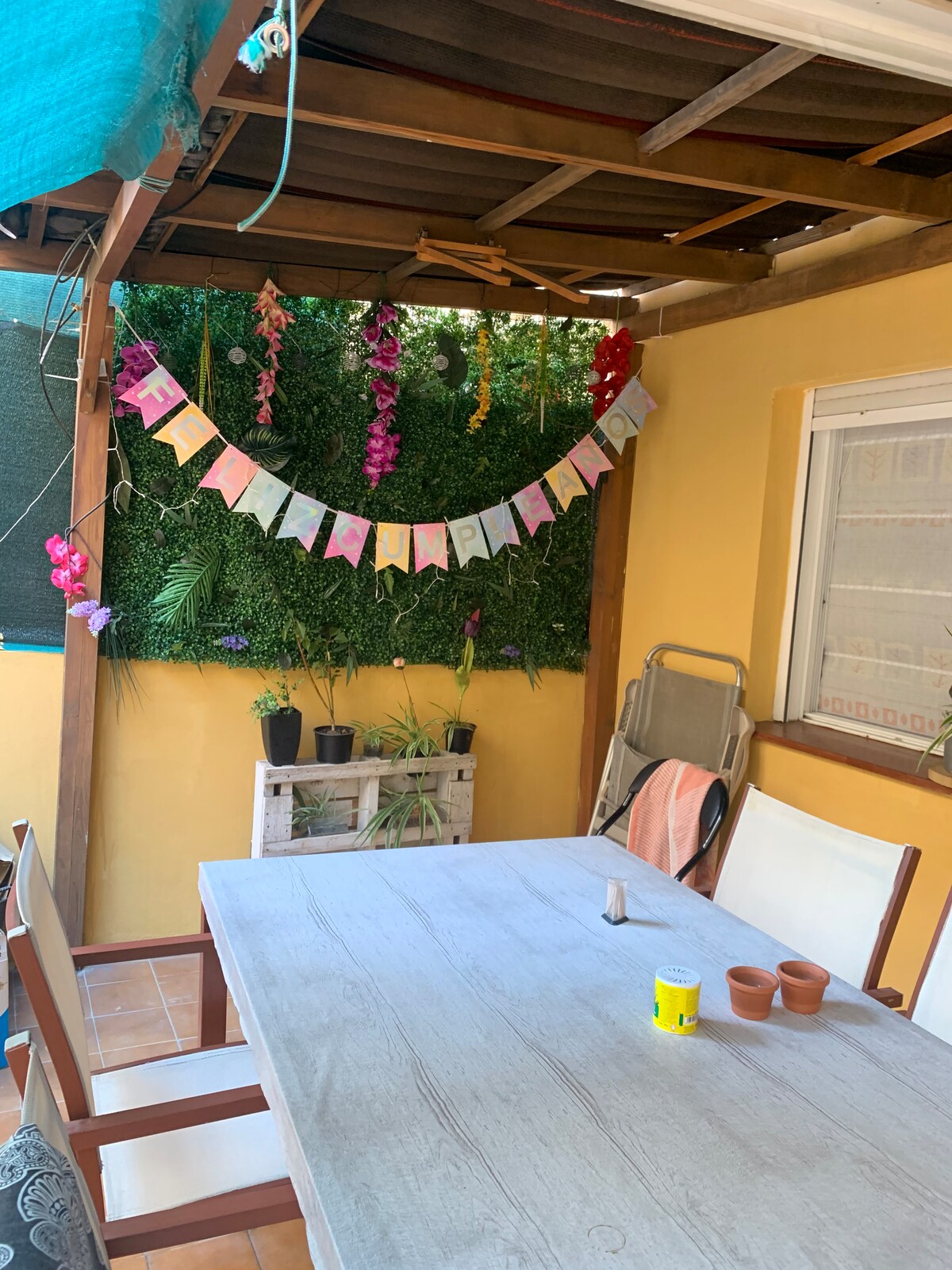 An outdoor terrace is presented with a wooden table surrounded by four chairs. Colorful decorations hang from the overhead structure, and potted plants are placed along the side walls. A backdrop of greenery adds a natural touch, contributing to a relaxed atmosphere.