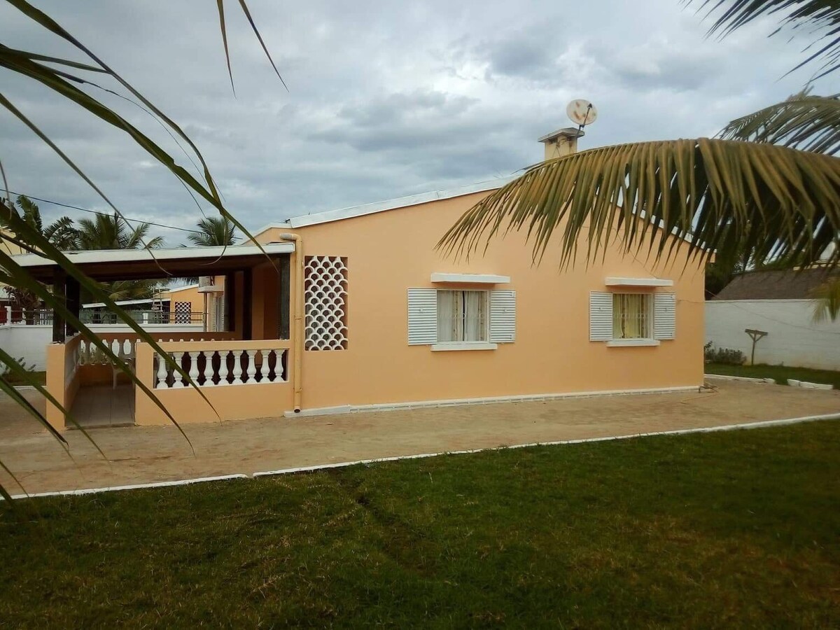 A single-story villa with an orange exterior is set against a cloudy sky. White shutters frame the windows, and a covered porch features decorative white railings. Green grass surrounds the property's edges, creating a harmonious outdoor space.