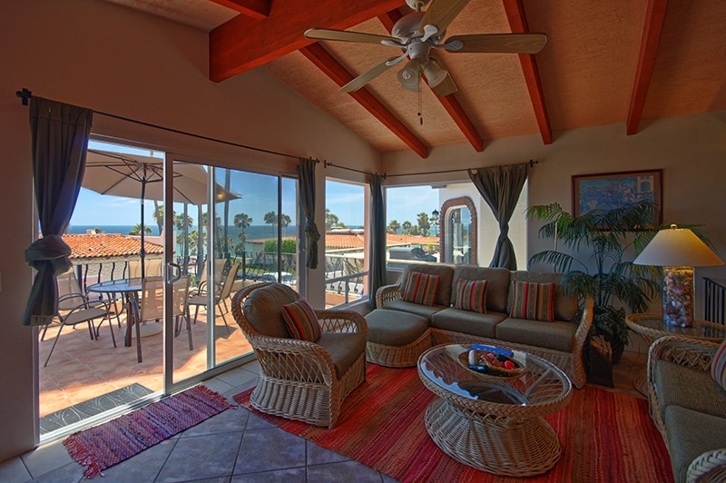 The spacious living room features large sliding glass doors that open to a balcony with ocean views. Natural light enhances the room, which is furnished with a comfortable seating arrangement and a decorative area rug. A ceiling fan hangs above for ventilation.
