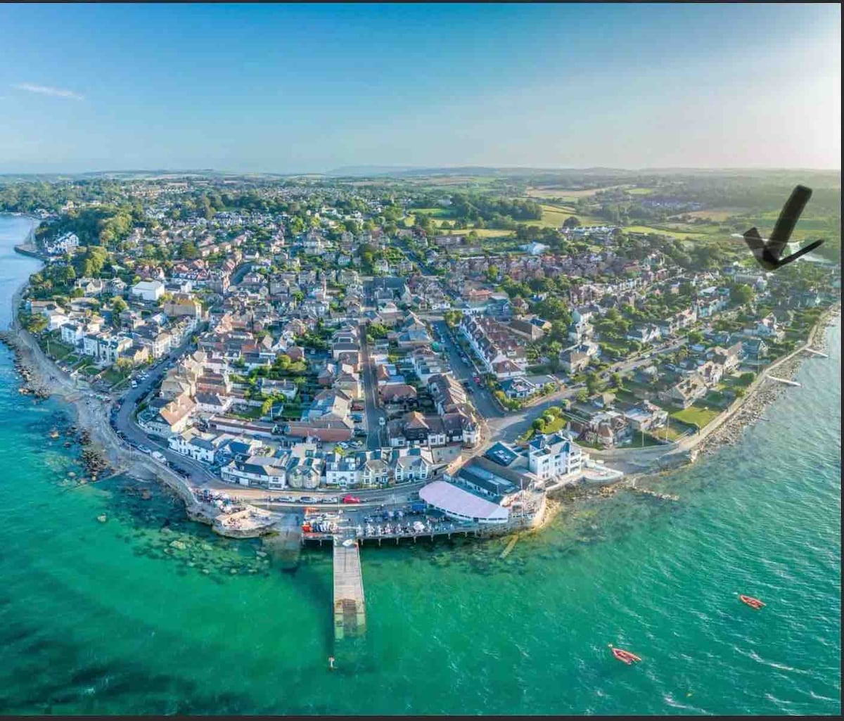 An aerial view showcases Seaview's charming village layout, with residential buildings lining the coastline. The clear blue waters surround the area, and a pier extends from the shore, providing a focal point for the bustling community. Green hills rise in the distance.